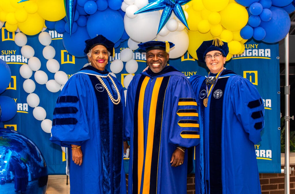 Group of SUNY Niagara employees smiling at the camera in black graduation robes