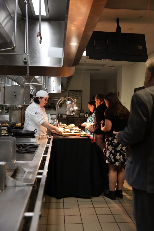 A chef in a white uniform serves food to a group of people standing in line inside a commercial kitchen with stainless steel equipment and overhead lighting.