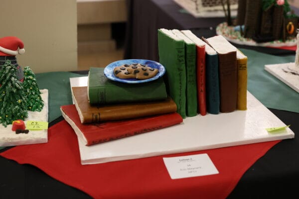 A cake designed to look like a stack of colorful books with a blue plate holding chocolate chip cookies on top, displayed on a white board with a red cloth underneath.
