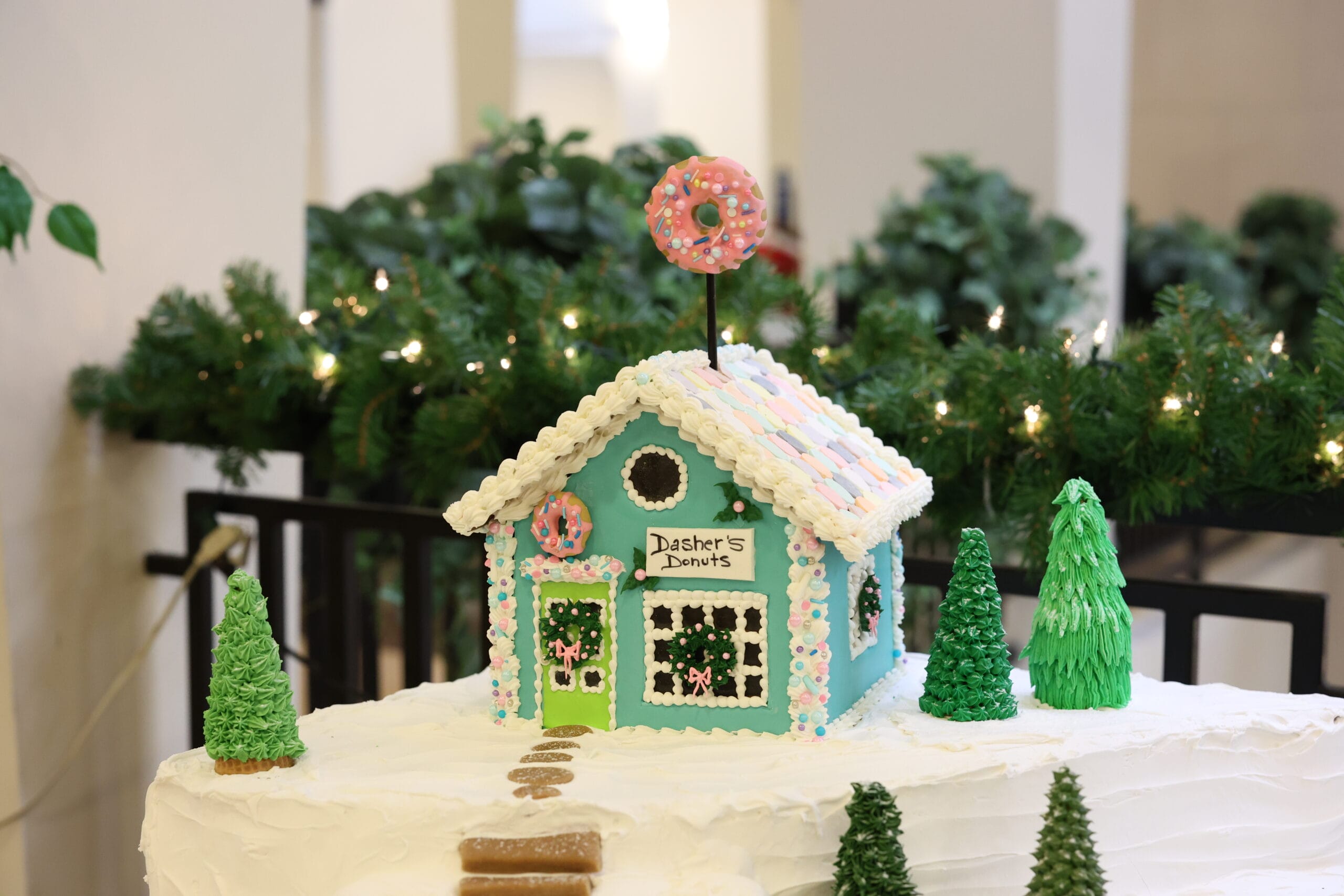 A decorated gingerbread house with a sign reading Dashers Donuts, candy details, and a pink donut on top sits on a snowy base, surrounded by green icing trees and festive greenery in the background.
