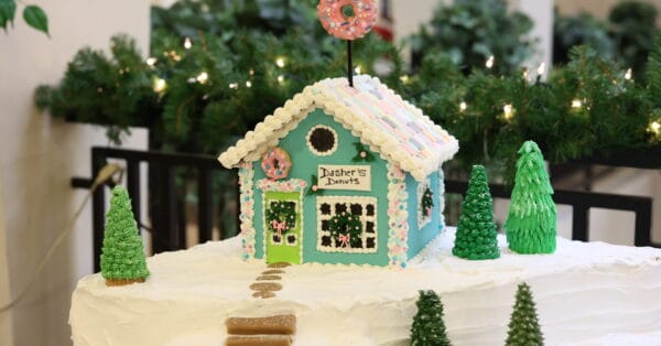A blue gingerbread house decorated with icing, labeled Dashers Donuts, sits on snowy white frosting with candy trees and a donut sign on the roof. Green garland is in the background.