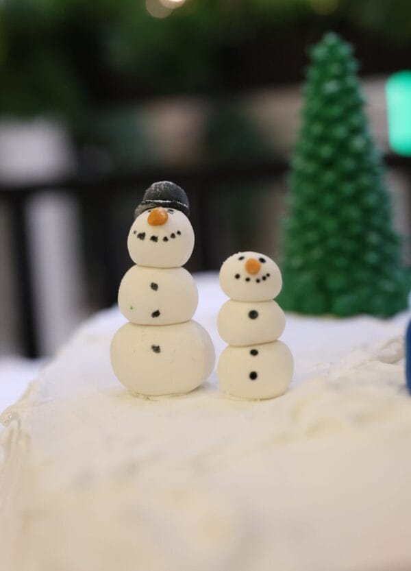 Two small snowman figurines made of white balls, one with a black hat, stand on a snowy surface with a green tree decoration in the background.