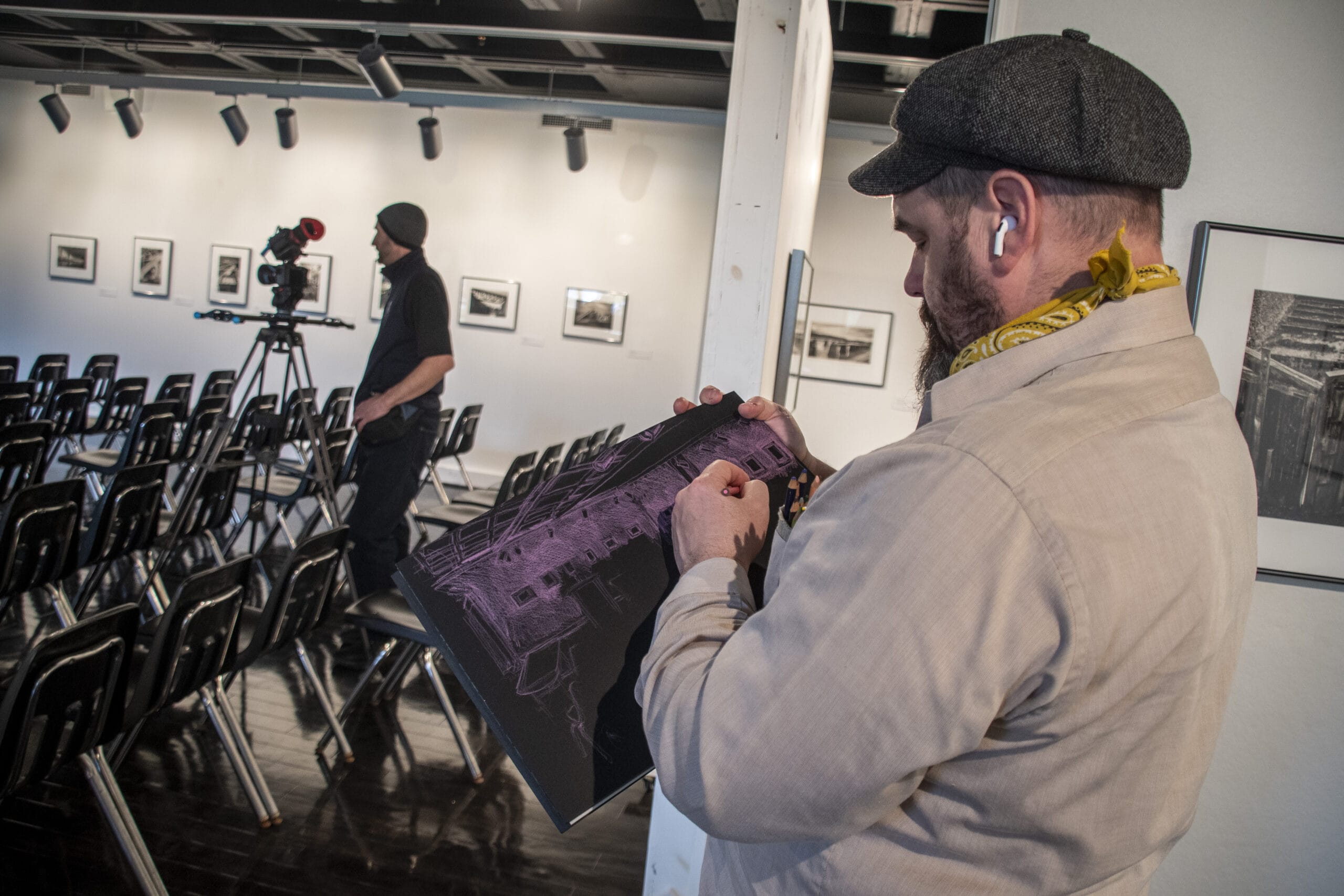 A man stands in an art gallery looking at a printed image while wearing earbuds and a cap. In the background, another person operates a camera in front of empty rows of chairs and framed photos on the walls.