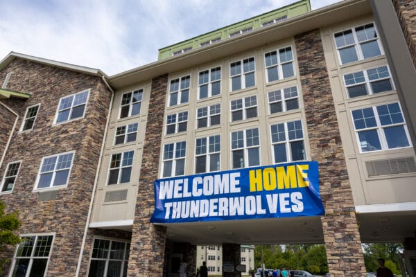 A large banner reading WELCOME HOME THUNDERWOLVES hangs on the exterior of a multi-story stone and beige building at the Student Housing Village at SUNY Niagara—apply now for the 2024-2025 academic year!.
