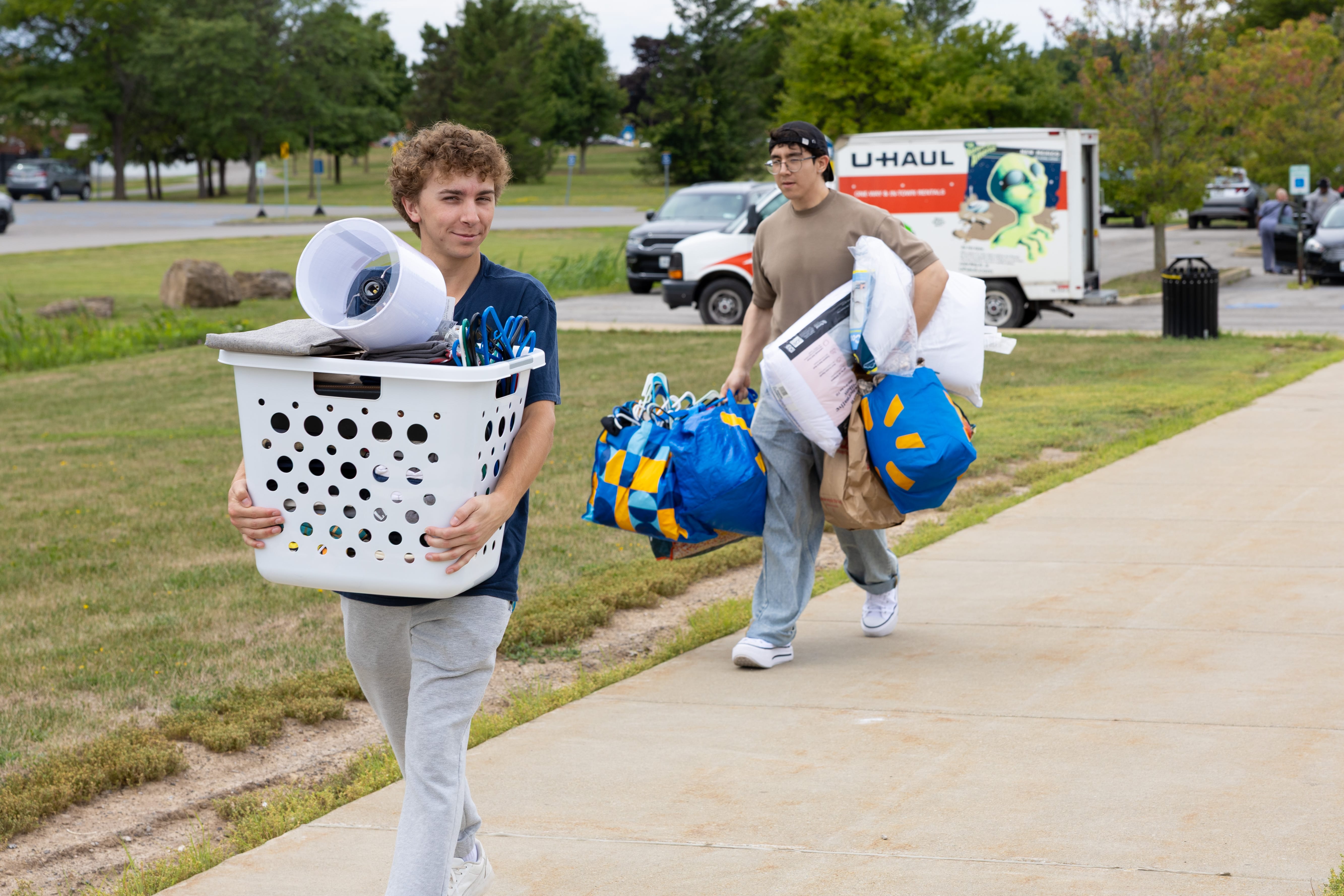 Two young men carry laundry baskets along a sidewalk near a U-Haul, suggesting moving day at a college campus—perhaps they're headed to Student Housing Village at SUNY Niagara. Apply now for the 2024-2025 academic year!.