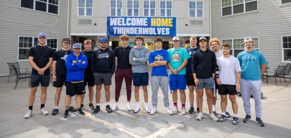 A group of 15 young men stand together outside a building with a Welcome Home Thunderwolves banner above the entrance at the Student Housing Village at SUNY Niagara. Apply now for the 2024-2025 academic year! They are smiling and casually dressed.