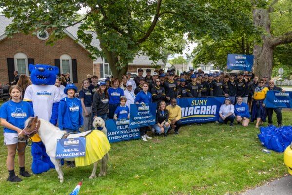 A large group of SUNY Niagara students, staff, and mascots gather outdoors holding signs for campus organizations, showcasing Campus Life at SUNY Niagara: Engaging Events and Leadership Opportunities for Student Development.
