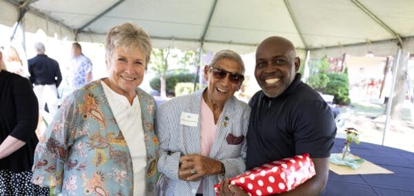 Three people smiling together under a tent at an outdoor event; one holds a red polka-dotted gift, another wears sunglasses and a striped jacket, and the third wears a patterned top. Tables and greenery can be seen in the background.