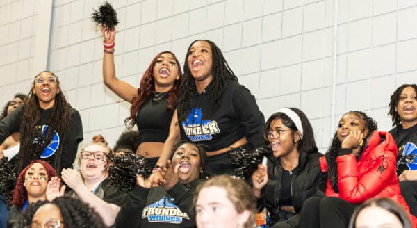 A group of enthusiastic young women cheer loudly in the stands of a gym, some holding pom-poms and wearing Thunder Wolves shirts, showing excitement and school spirit.