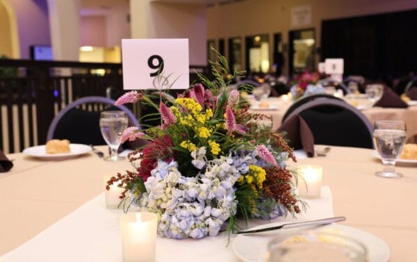 A banquet table with a floral centerpiece, candles, water glasses, and plates. A card with the number 9 stands in the centerpiece. The background shows other tables set for an event.