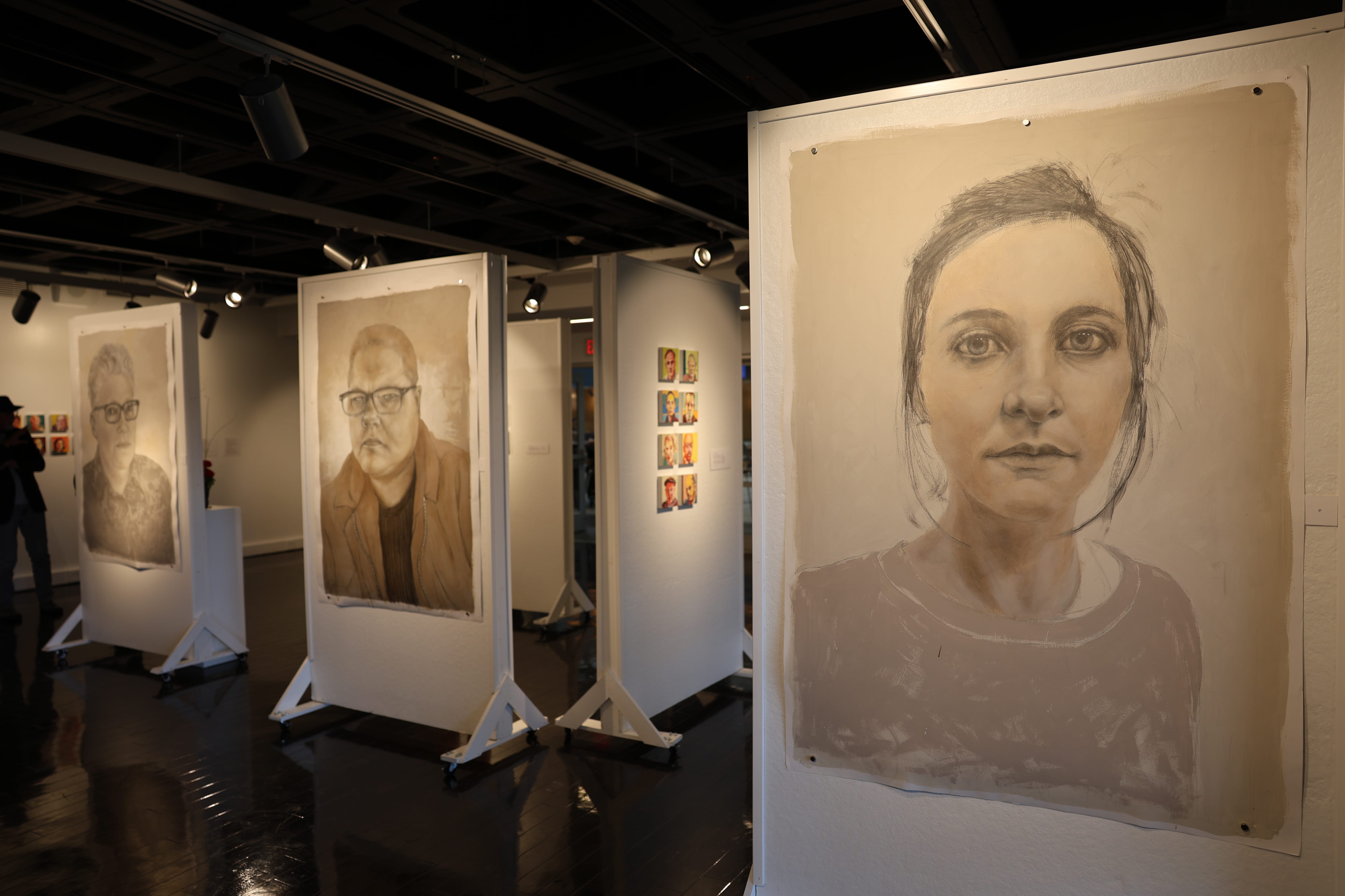 Large grayscale portraits of people are displayed on wheeled white panels in an art gallery with dark floors and ceiling. Smaller colorful artworks are visible in the background. The space is softly lit.