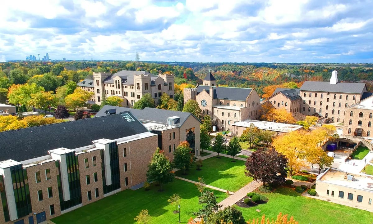 Aerial view of a university campus with stone buildings, green lawns, and autumn trees; the city skyline rises in the distance under a partly cloudy sky. Discover SUNY Niagara: Two Locations, where one vibrant community thrives amid stunning scenery.