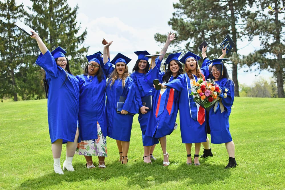 Seven graduates in blue caps and gowns stand on grass, smiling and cheering with arms raised. One holds a bouquet of flowers. Trees and a bright sky are in the background, celebrating success—apply to SUNY Niagara to start your journey.