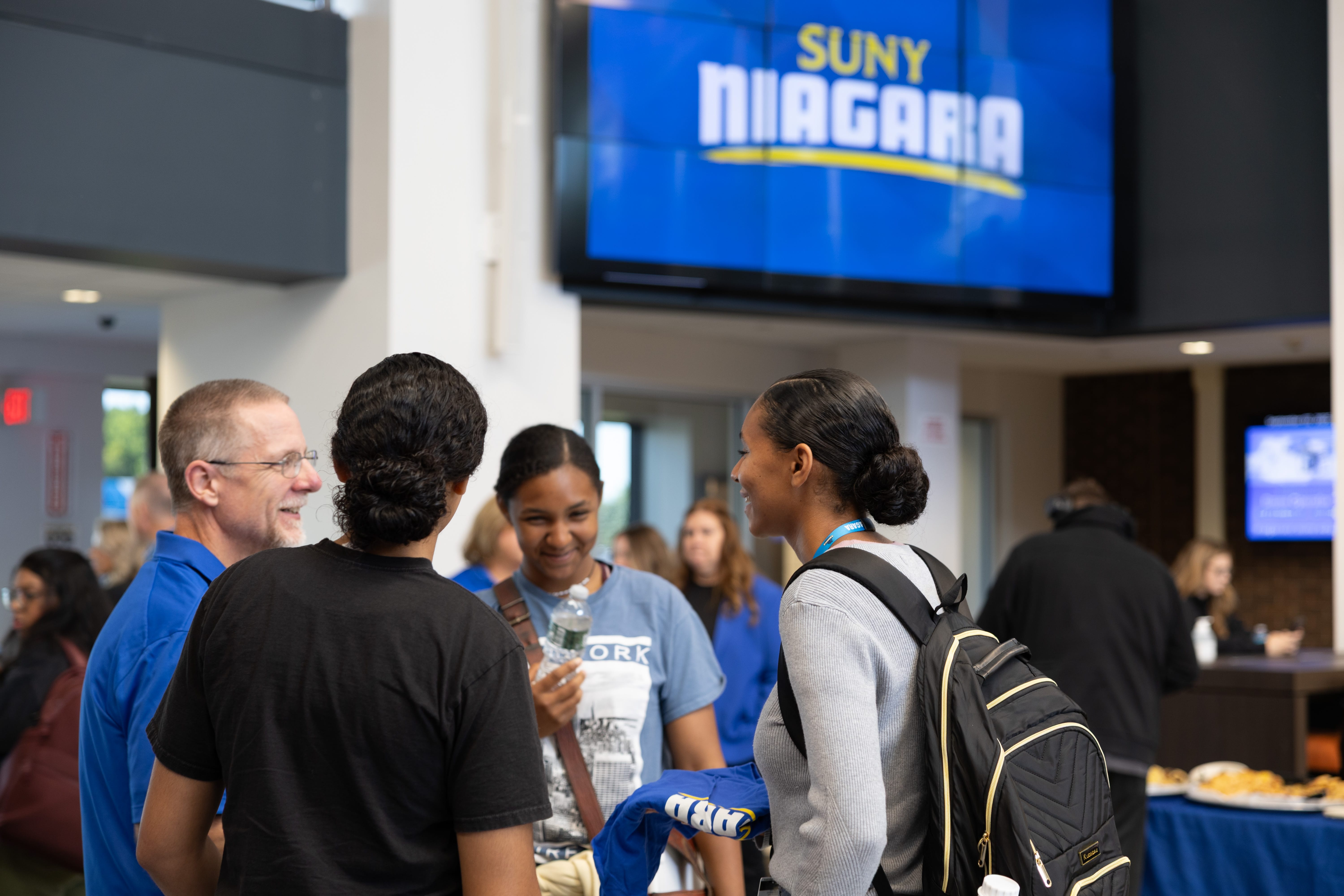 A group of people smiling and talking inside a building, with a large screen in the background displaying SUNY NIAGARA. Other people are visible in the background near tables with food.