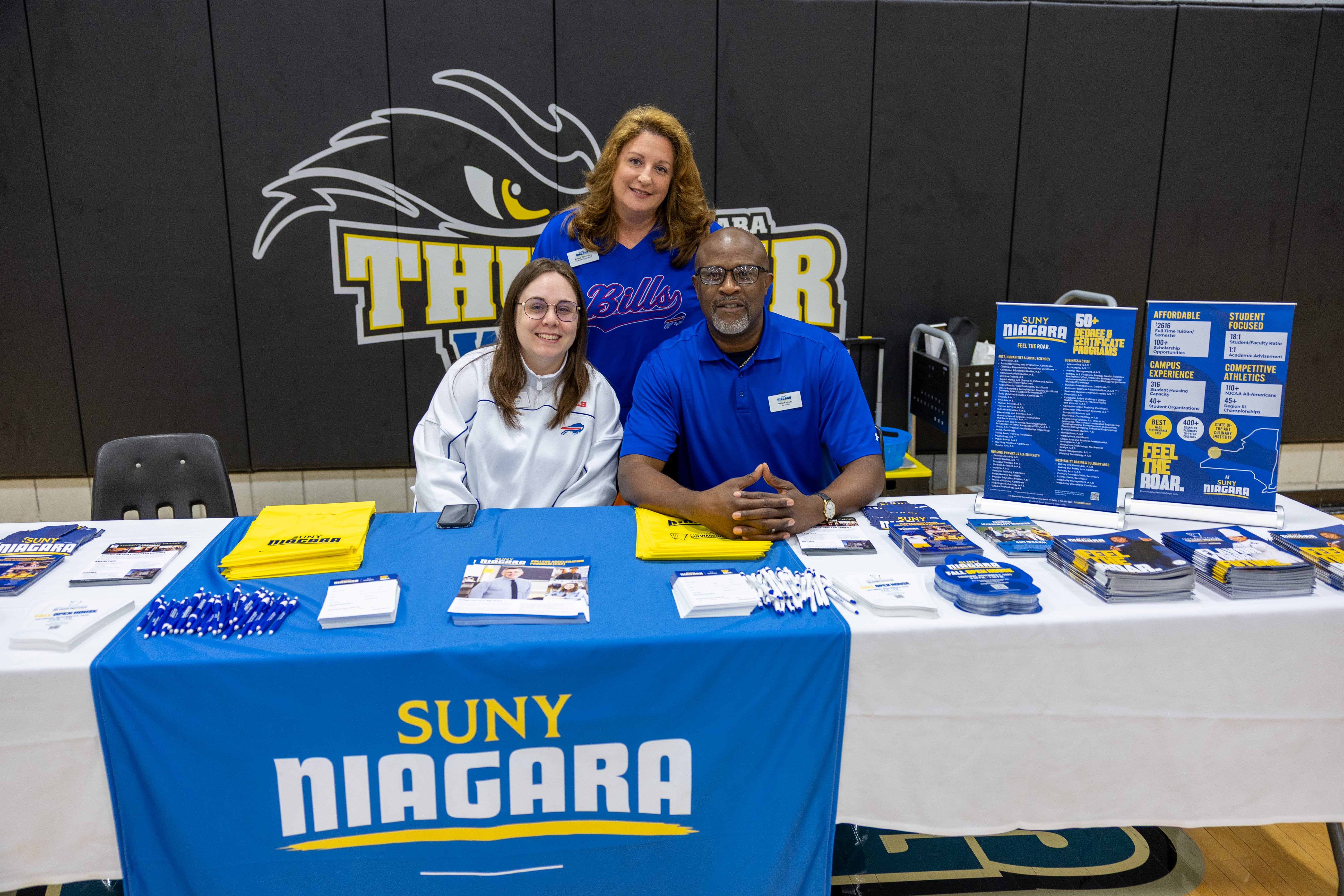 Three smiling people stand and sit behind a SUNY Niagara information table with brochures, pens, and promotional items. The table is in a gym, with a Thunder Wolves logo on the wall behind them.