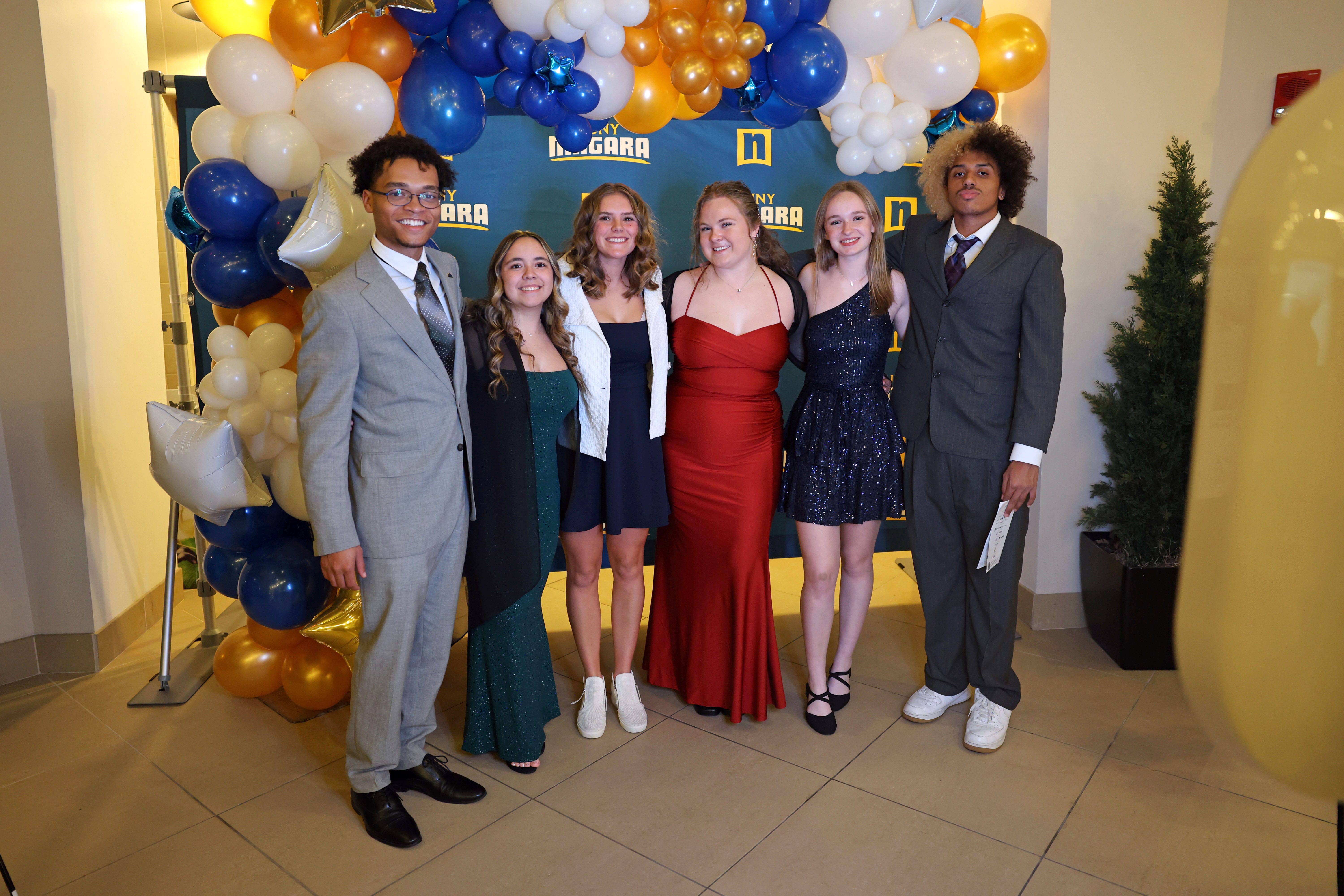 Six young people dressed in formal wear stand together and smile in front of a Niagara-themed backdrop, surrounded by white, blue, and gold balloons in a well-lit indoor space.