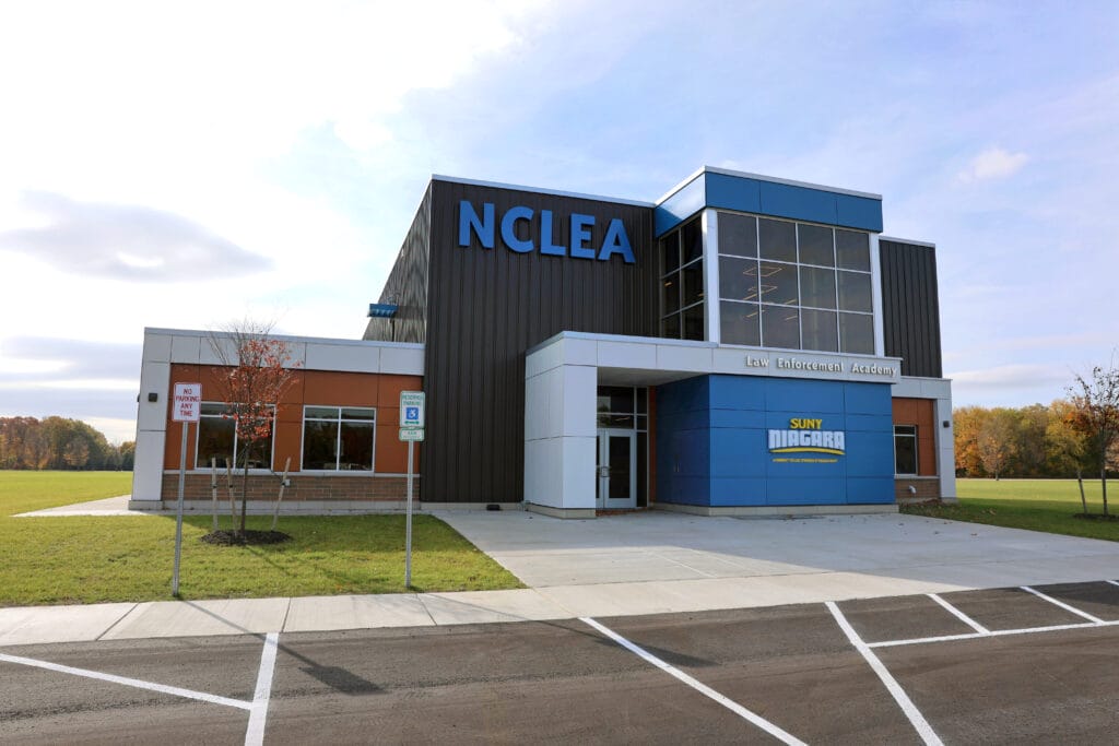 Modern building with “NCLEA” in large blue letters on the facade, labeled as the Law Enforcement Academy of SUNY Niagara; surrounded by grass, parking spaces, and a clear sky.