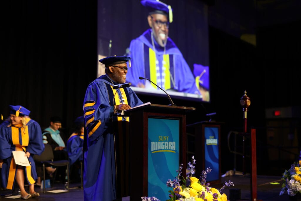 A speaker in academic regalia addresses an audience at a podium labeled SUNY Niagara during a graduation ceremony, with a large screen behind him showing his image. Other faculty are seated on stage.