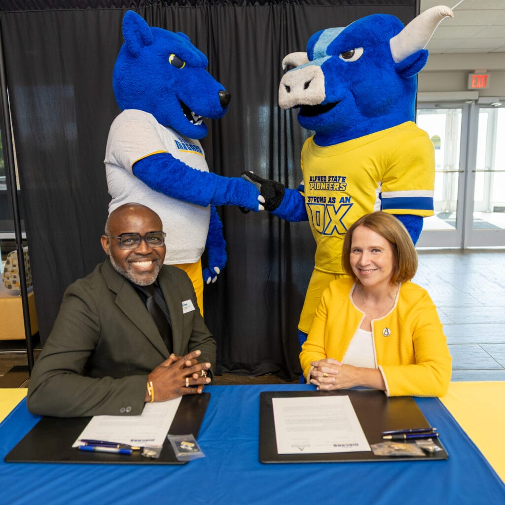 Two college mascots, a blue wolf and a blue ox, shake hands behind a table where a smiling man in a dark suit and a smiling woman in yellow sit with documents and pens, suggesting a partnership signing event.