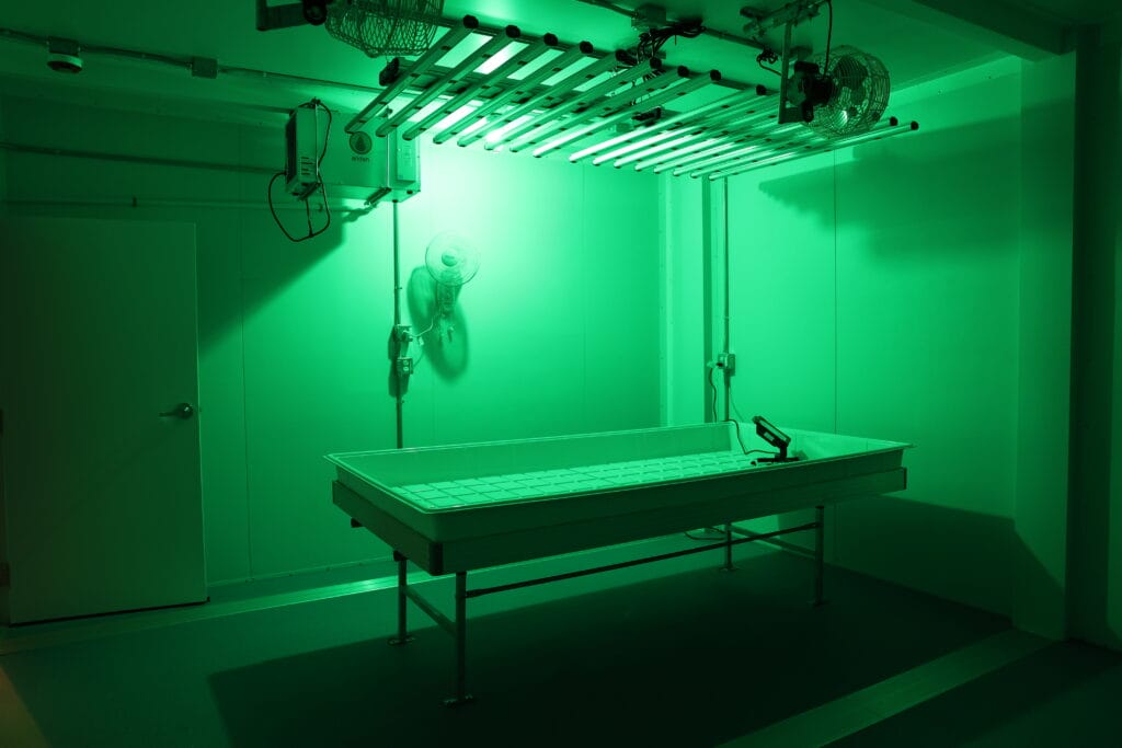 A white, rectangular table stands in the center of an empty room lit by green lights from the ceiling. A mounted fan and control panels are visible on the wall, and a closed door is in the background.