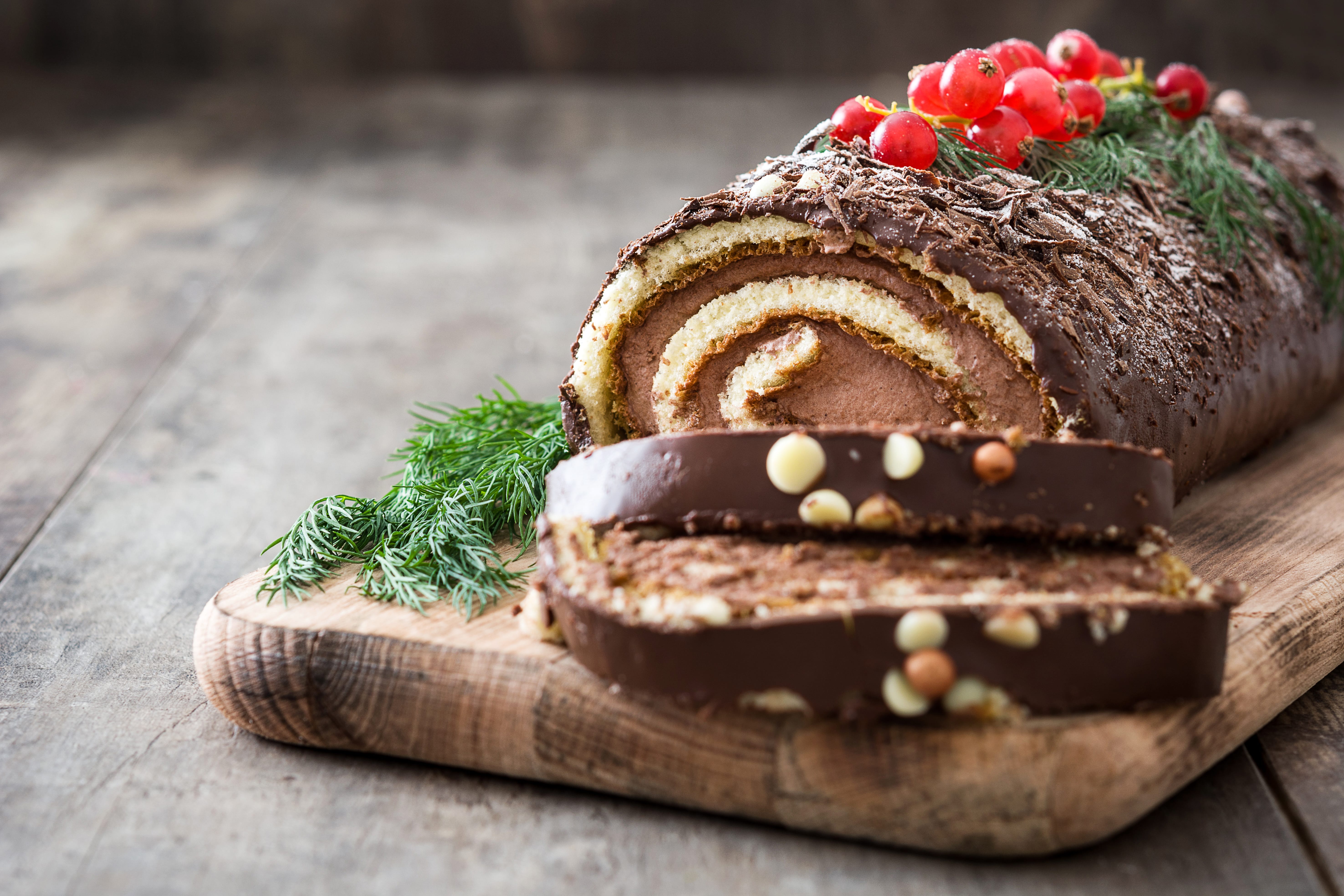A chocolate yule log cake with a spiral filling, topped with red berries, chocolate shavings, and green herbs, sits on a wooden board with two slices cut at the end.