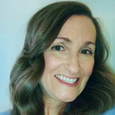 A woman with long, wavy brown hair smiles at the camera. She is wearing a light blue blazer over a dark top, with a light-colored background behind her.