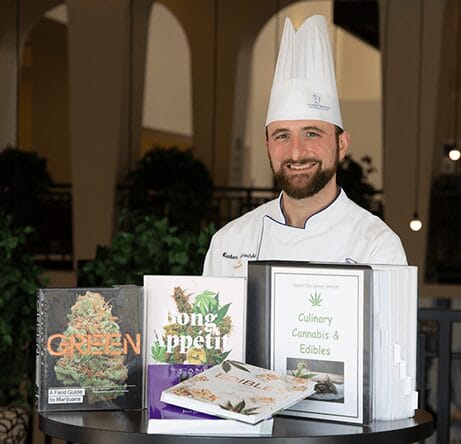 A smiling chef in a white uniform and hat stands behind a table displaying several cannabis-themed cookbooks, including titles like Bong Appétit and Culinary Cannabis & Edibles, in a well-lit indoor space.