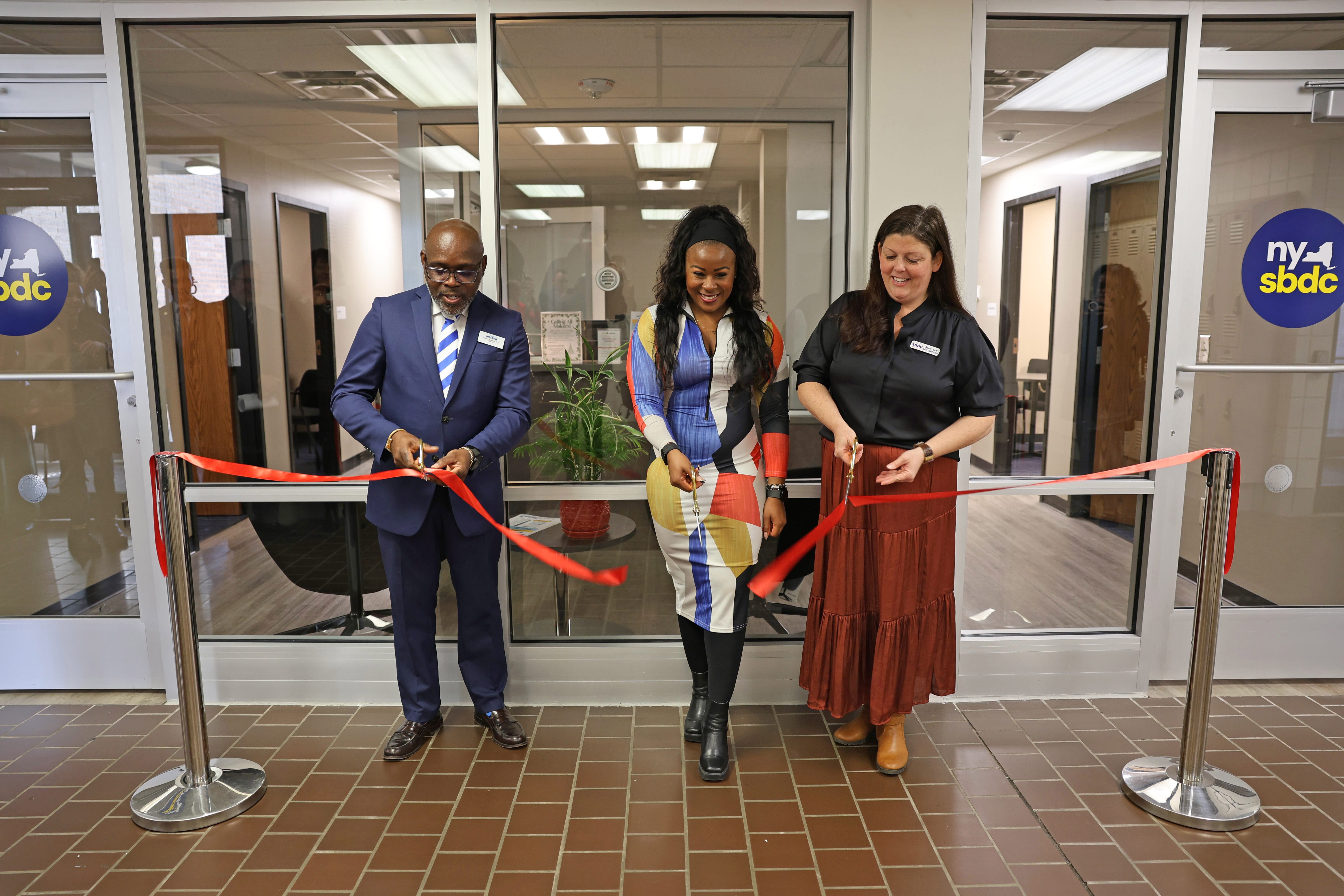 Three people stand behind a red ribbon at an indoor ribbon-cutting ceremony. The woman in the center cuts the ribbon with large scissors while the two others smile nearby. Offices with ny sbdc signs are visible in the background.