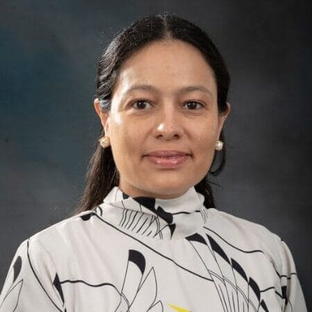 A woman with long dark hair, wearing a white blouse with crane bird designs and pearl earrings, stands in front of a dark, neutral background, looking at the camera with a slight smile.