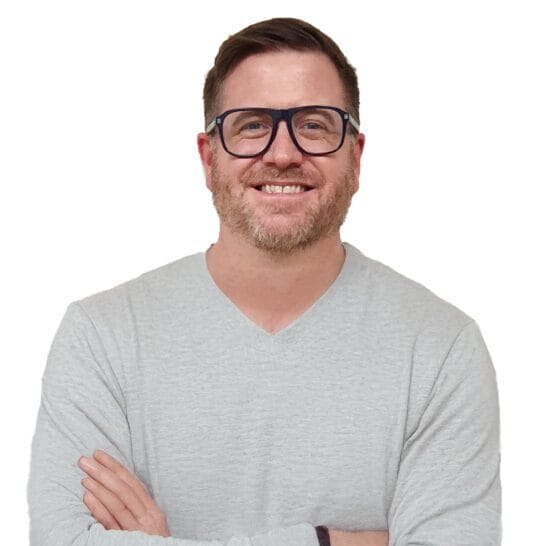 A man with short brown hair, a beard, and glasses is smiling with his arms crossed. He is wearing a light gray long-sleeve shirt and a dark wristband, standing against a plain white background.