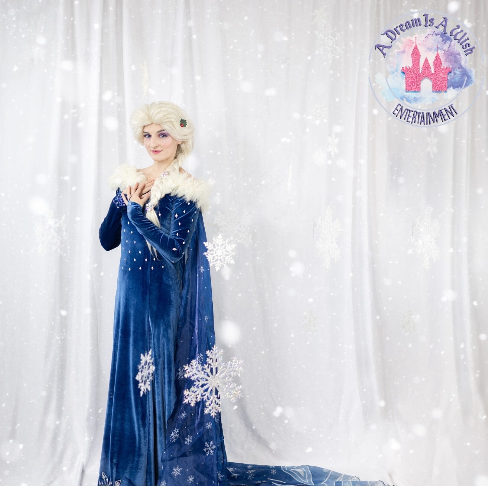 A person dressed as a snow queen in a long blue velvet gown with white fur trim stands against a snowy, winter-themed backdrop with snowflakes and the logo A Dream Is A Wish Entertainment in the corner.