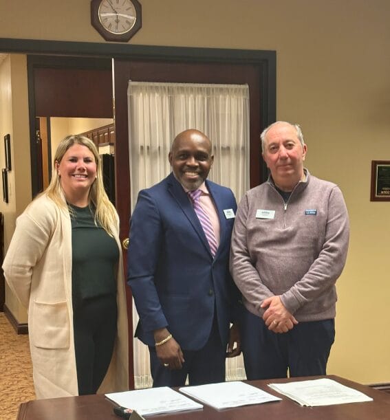 Three people stand indoors behind a table with documents. The woman on the left wears a light coat, the man in the center wears a blue suit, and the man on the right wears a mauve sweater. A clock and plaques are visible on the wall.
