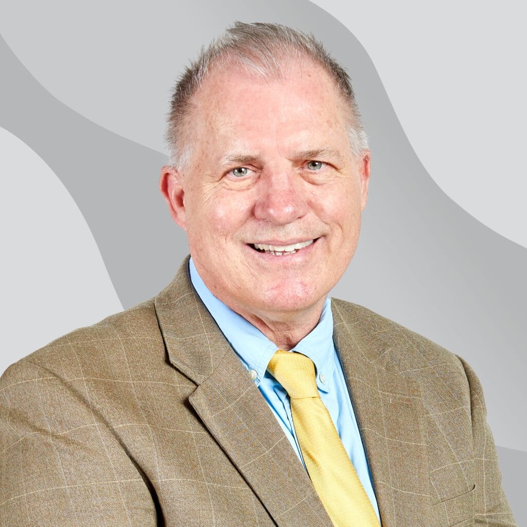 A smiling older man with short gray hair wears a beige plaid suit jacket, light blue shirt, and yellow tie, posing in front of a gray background with wavy patterns.