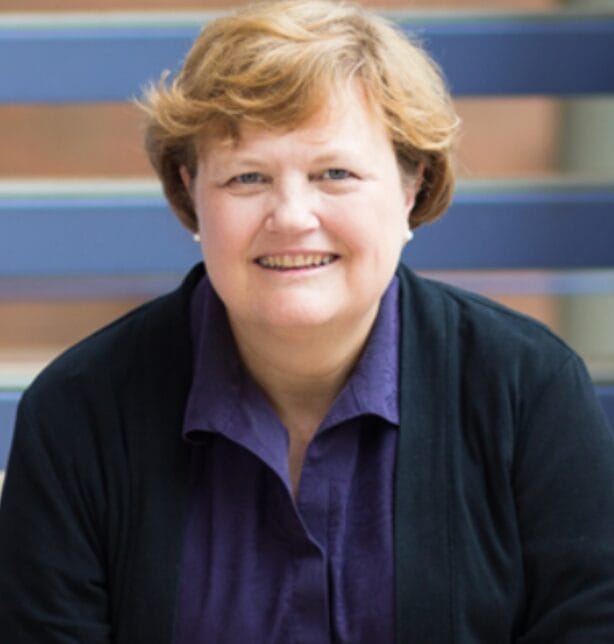 A smiling woman with short, light brown hair wearing a purple shirt and a dark cardigan, sitting in front of blue horizontal railings.