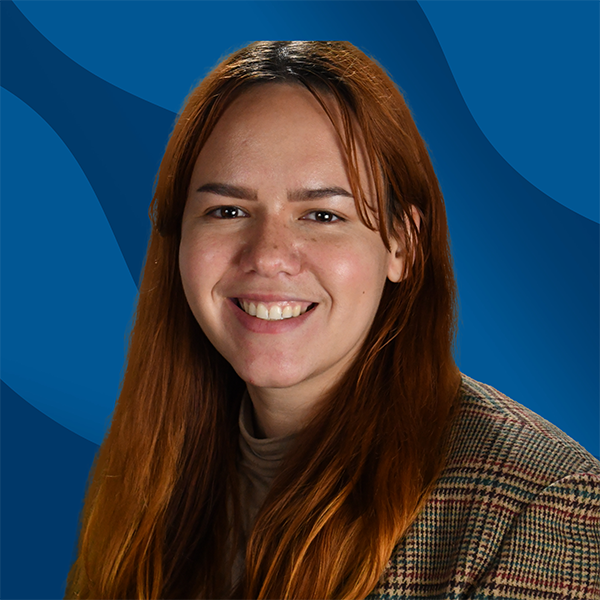 A woman with long reddish-brown hair smiles at the camera. She is wearing a brown plaid jacket and beige top, standing before a blue wavy background—representing the supportive spirit of the Student Success Center: Your Hub for Success Coaching and more.