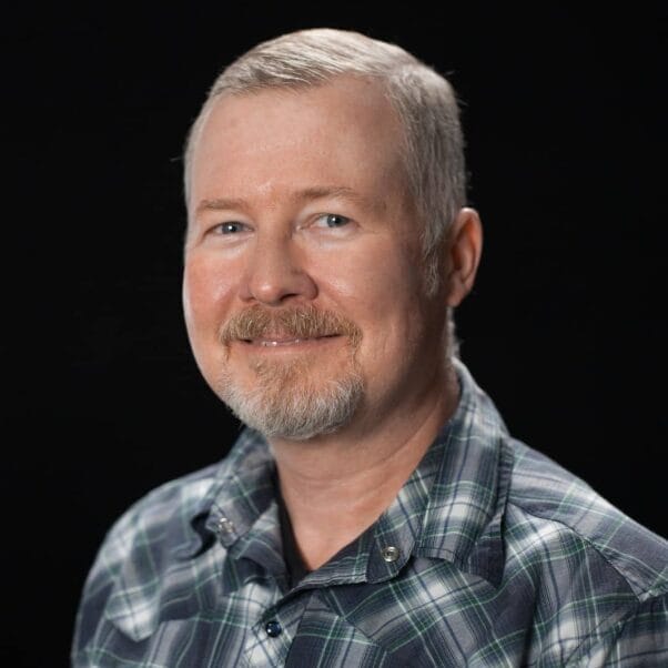 A middle-aged man with short gray hair and a trimmed beard is smiling. He is wearing a green and blue plaid button-up shirt and is posed against a plain black background.