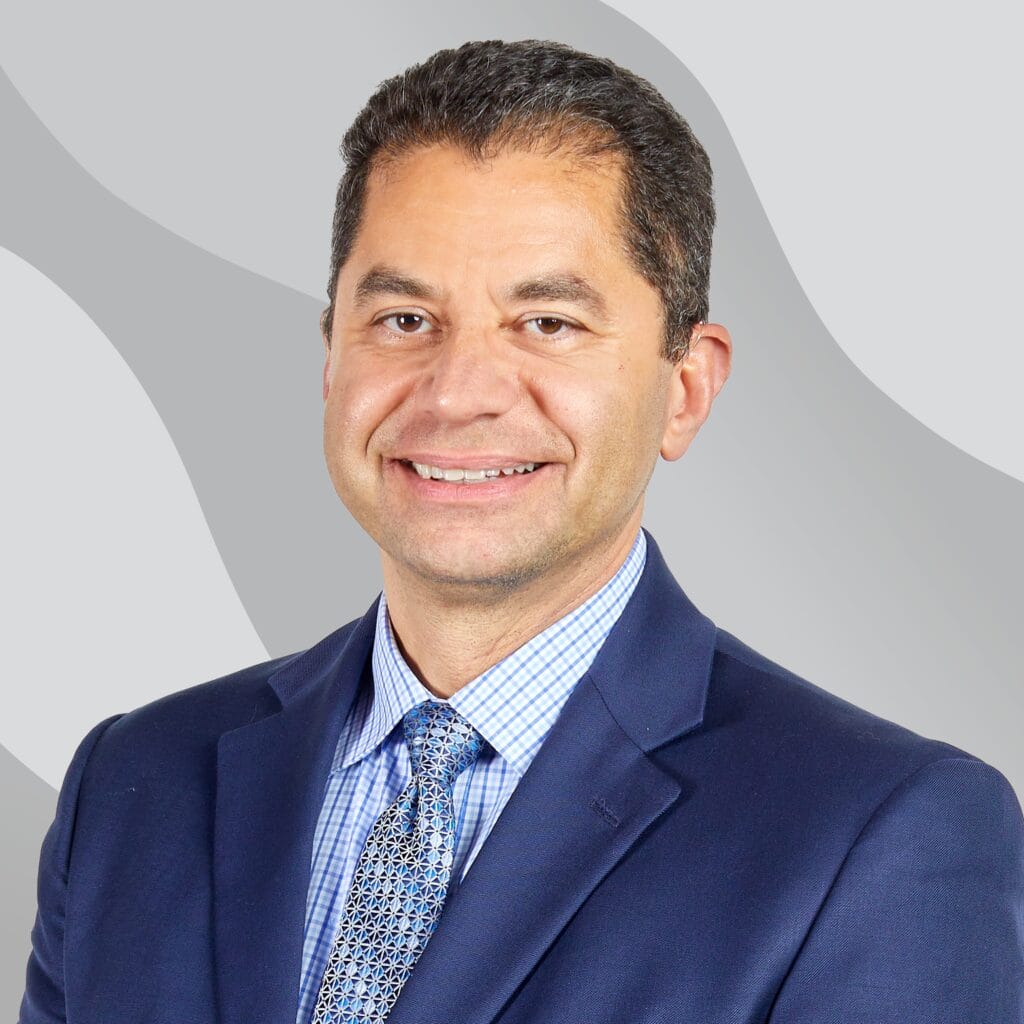 A man wearing a blue suit, patterned tie, and checkered shirt smiles at the camera against a gray background with abstract wavy shapes.