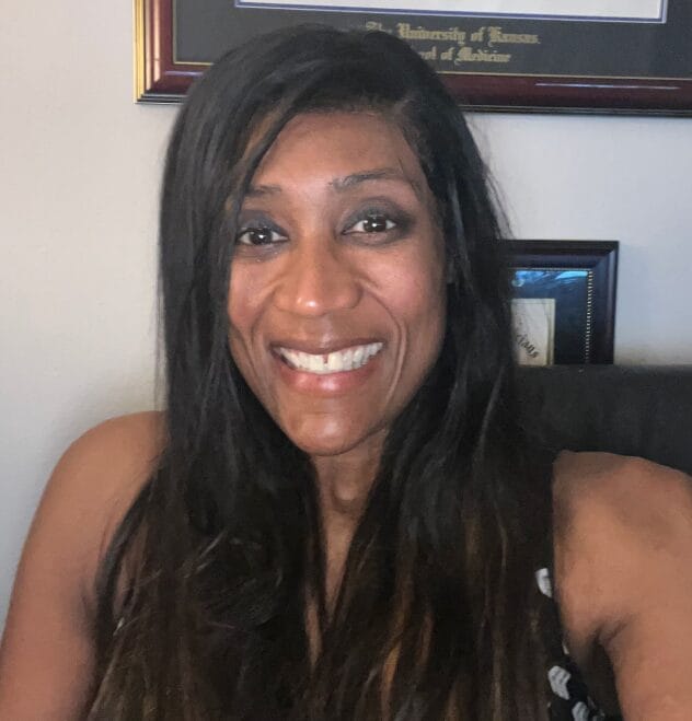 A woman with long dark hair smiles at the camera in an indoor setting. She is wearing a sleeveless top, and framed certificates or diplomas hang on the wall behind her.
