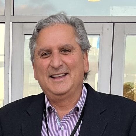 A smiling middle-aged man with gray hair, wearing a black blazer and a striped shirt, stands in front of glass doors.