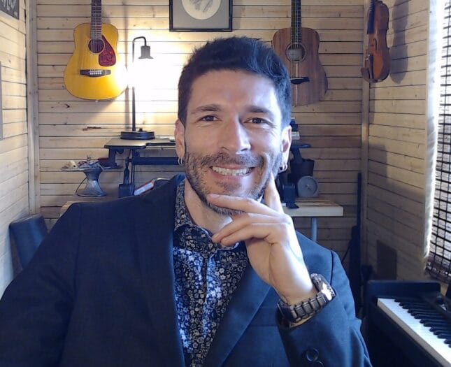 A man in a dark suit smiles at the camera, sitting in a wooden room decorated with hanging guitars, a piano, and a window with blinds letting in sunlight.