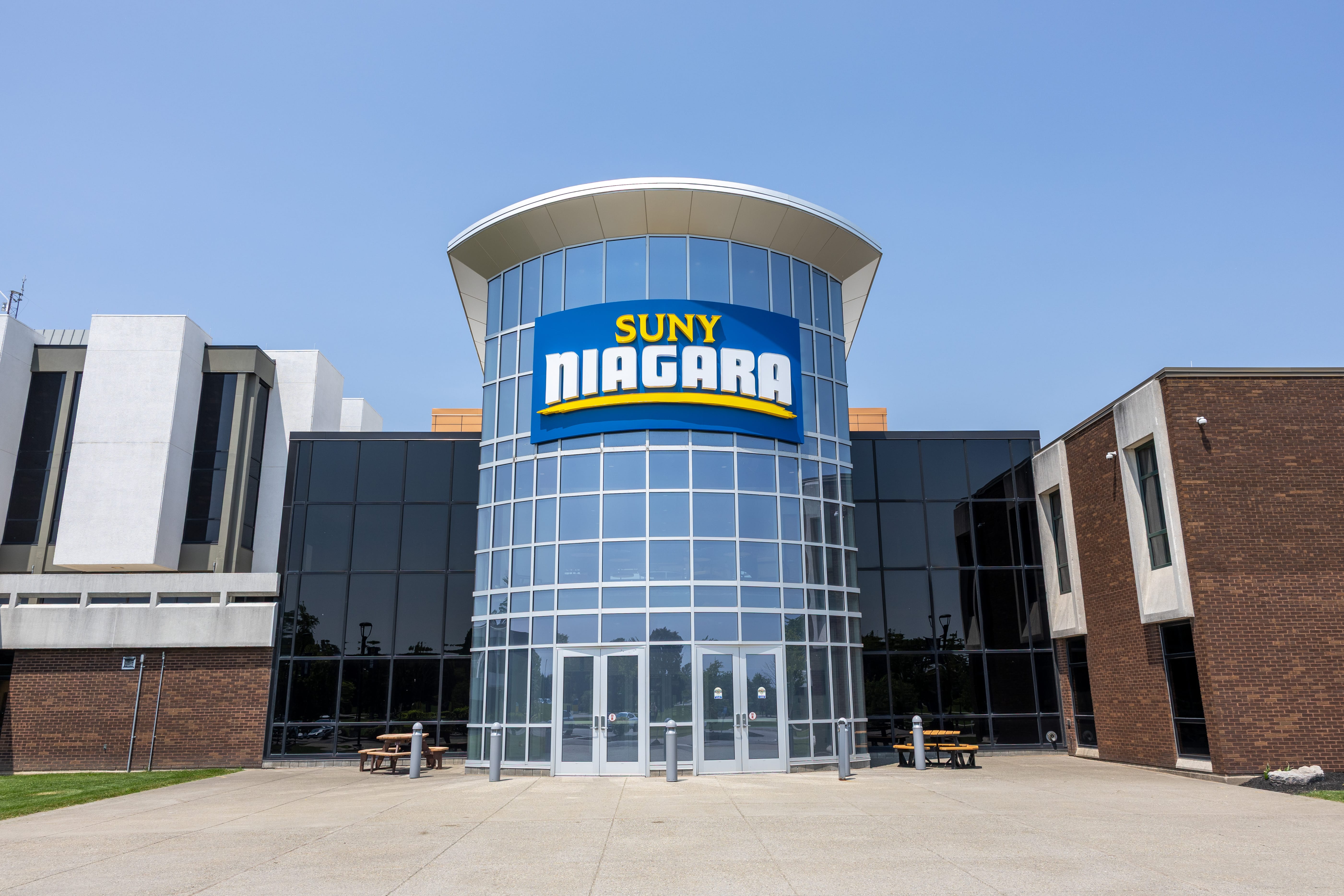 The entrance of a modern building with large glass windows and a sign reading SUNY NIAGARA above the doors, with clear skies and two benches outside.