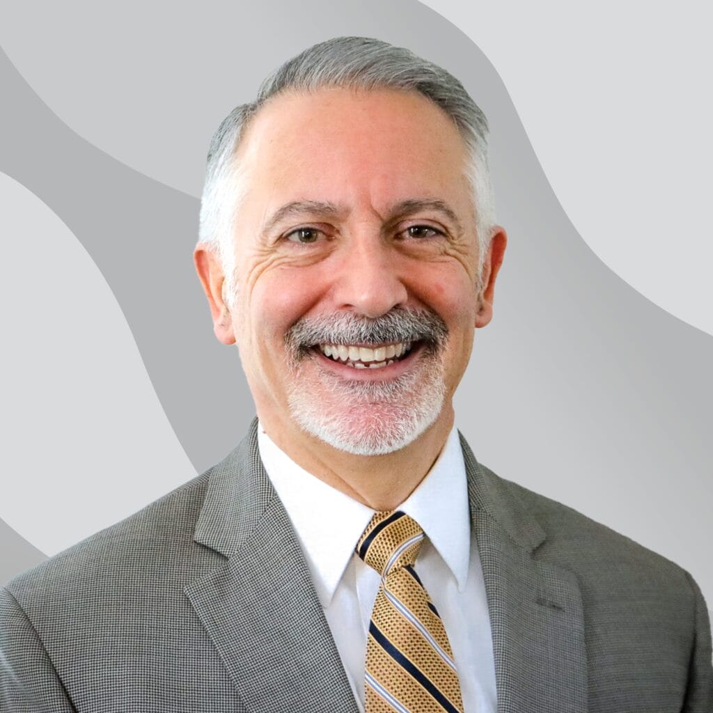 A middle-aged man with gray hair and a mustache, wearing a gray suit, white shirt, and striped tie, smiles at the camera against a light gray wavy-patterned background.