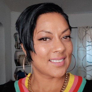 A woman with short black hair smiles at the camera. She is wearing a black top with a colorful striped V-neck, large hoop earrings, and a gold chain necklace. Shelves and a window with curtains are in the background.