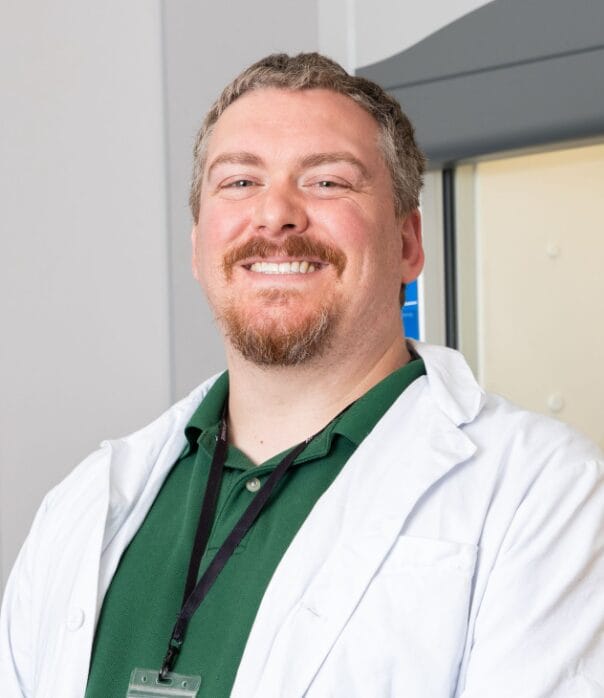 A man with short light brown hair, beard, and mustache smiles while wearing a white lab coat over a green shirt and a black lanyard. He stands in an indoor setting with a light-colored background.