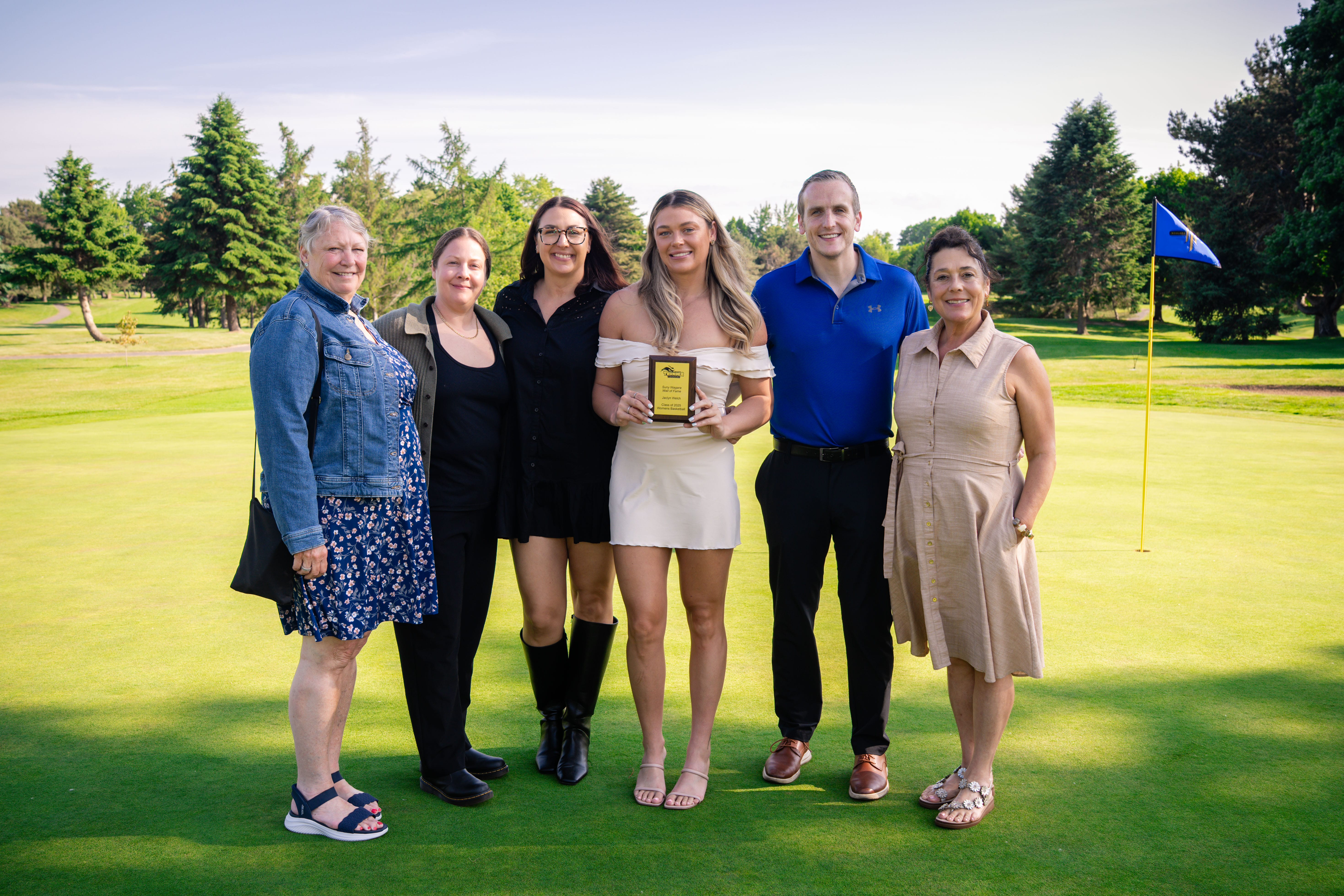 Six people stand together smiling on a golf course green. One woman in the center holds a trophy. Trees and a golf flag are visible in the background under a clear sky.