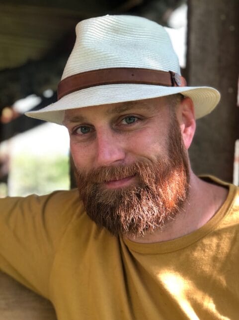 A bearded man wearing a white fedora hat with a brown band and a mustard yellow shirt smiles softly, sitting outdoors in natural sunlight.