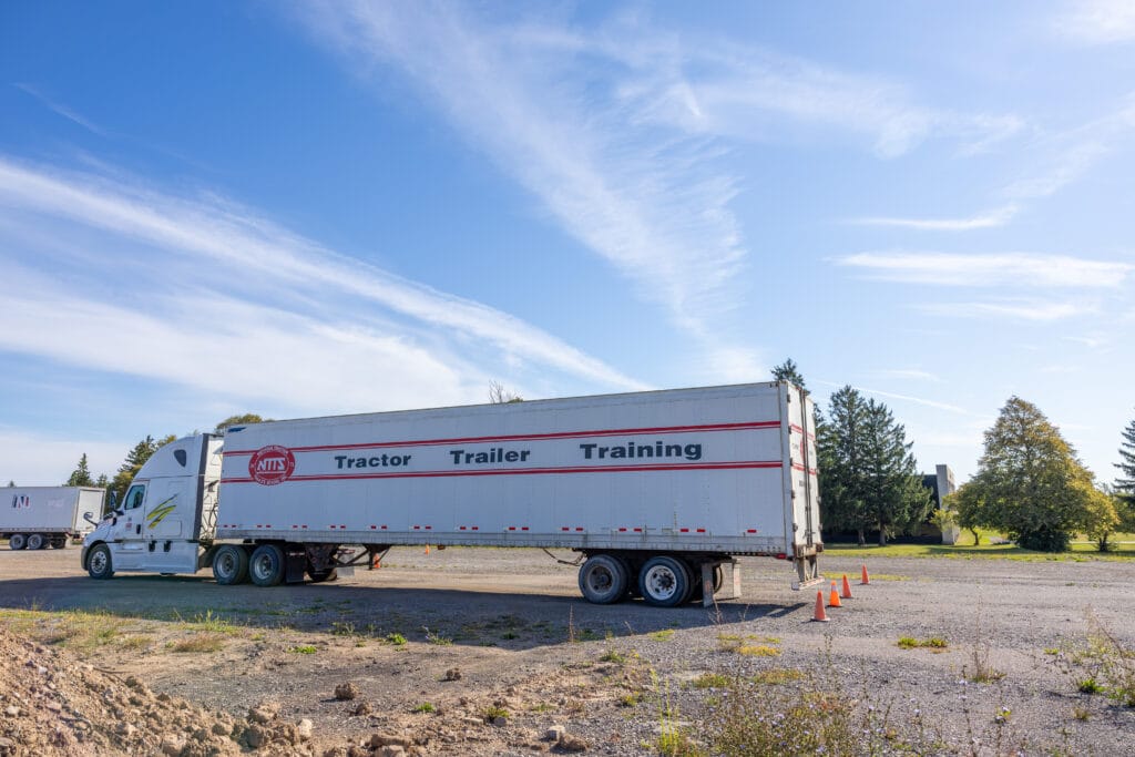 A white semi-truck with a trailer labeled “Tractor Trailer Training” is parked on a gravel lot, with orange cones nearby and trees and blue sky in the background.
