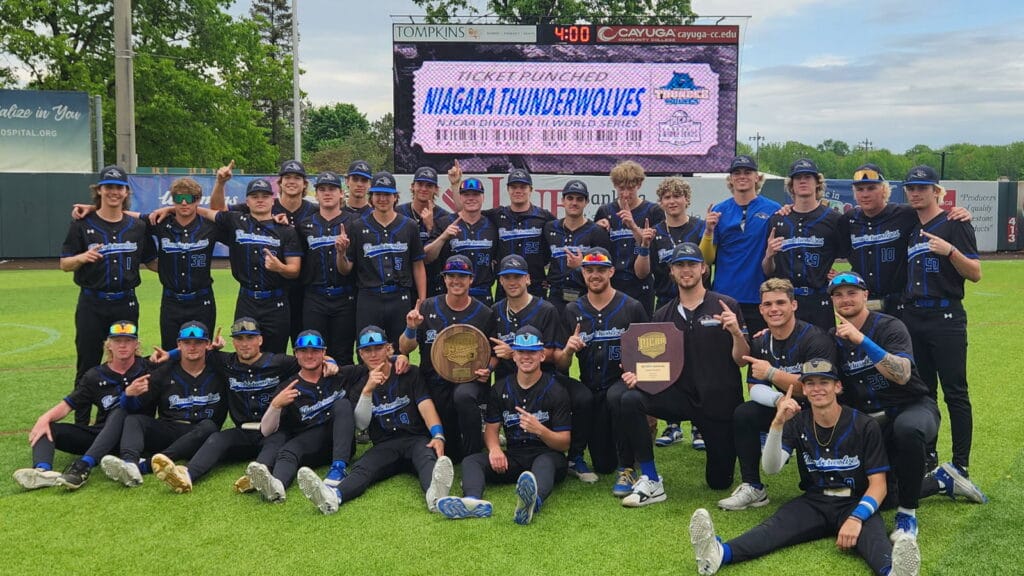 The Niagara Thunderwolves baseball team poses on a baseball field, smiling and holding up number one signs, with two large plaques and a scoreboard in the background reading Ticket Punched: Niagara Thunderwolves.