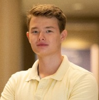 A young man with short light brown hair wearing a pale yellow polo shirt stands indoors with his arms crossed, looking confidently at the camera. The background is softly blurred.