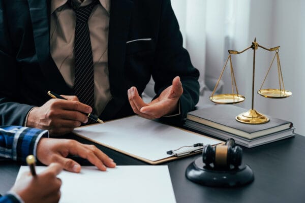 Two people sit at a desk with legal documents. One in a suit gestures while holding a pen. Scales of justice, a gavel, and law books are on the desk, suggesting a Workforce Development at SUNY Niagara legal consultation or meeting.